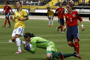 Selección Femenina de Colombia en partido contra Brasil, Copa América 2014, las colombianas visten la camiseta roja y la pantaloneta azul y Brasil con camiseta amarilla y pantaloneta blanca, la arquero Sandra Sepúlveda ataja el balón de frente y la fotografía muestra un congelado mientras cae al piso.